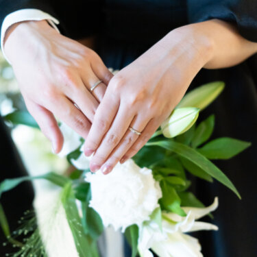 白いユリのブーケの上で、新郎新婦が指輪を着けた手を重ねてハートの形を作る手元のアップ。黒×黒コーデの前撮りで残したい指輪のカット