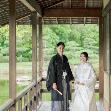 木の回廊の真ん中に並んで立ち、扇子を手に微笑む紋付袴の新郎と白無垢の花嫁。奥行きのある構図の和装前撮り