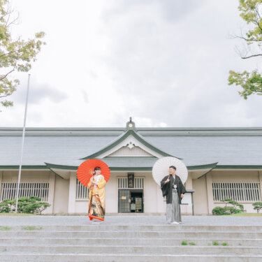 和風建築の前の階段の上で、赤い和傘の花嫁と白い和傘の新郎が離れて立つ構図。広がりのある和装ロケーションフォト