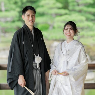 松の緑を背にして並び、扇子を持って笑顔を見せる新郎と白無垢の花嫁。大仙公園日本庭園で撮る結婚写真