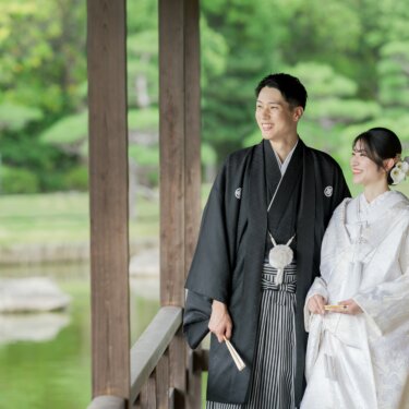 回廊の柱の間から、池のほとりに並んで立つ新郎新婦を写した構図。松の緑がさわやかな夏の和装フォトウェディング