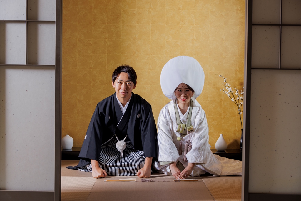 金箔の壁を背に、白無垢の綿帽子姿の花嫁様と黒紋付の花婿様が畳に座って微笑んでいる和室の写真