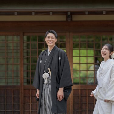 格子戸の前で向かい合い、大笑いする紋付袴の新郎と白無垢の花嫁。古い木造建築で撮るナチュラルな和装前撮り