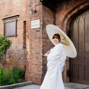 レンガ造りの教会の扉の前で、白い和傘をさしてほほえむ白無垢の花嫁。川口基督教会の外観で撮る花嫁ソロの和装前撮り