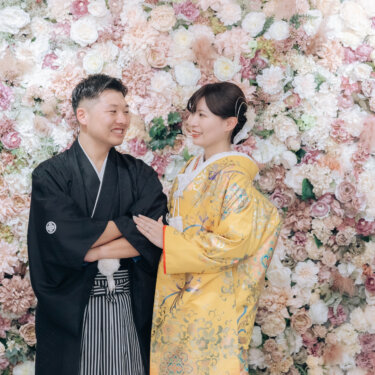 ピンクのバラのフラワーウォールの前で寄り添う、腕を組んだ新郎と色打掛の花嫁。心斎橋店のスタジオで撮る和装前撮り