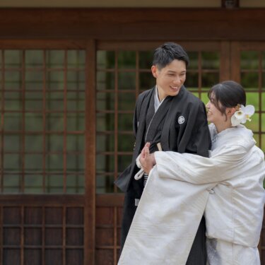 格子戸の前で新郎に抱きついて笑う白無垢の花嫁。照れる新郎との仲の良さが伝わる和装前撮りのハグショット