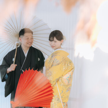 白い番傘と赤い扇子を手に寄り添う紋付袴の新郎と色打掛の花嫁。淡い光に包まれたスタジオの和装フォトウェディング
