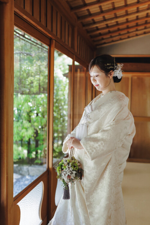 木の縁側の窓辺でブーケを手に目を伏せる白無垢の花嫁。生花の髪飾りを合わせた洋髪の和装前撮り（NS102）