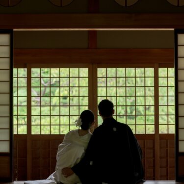 丸い欄間のある和室で、格子戸越しの庭を眺めて並んで座る白無垢の花嫁と紋付の新郎の後ろ姿の結婚写真