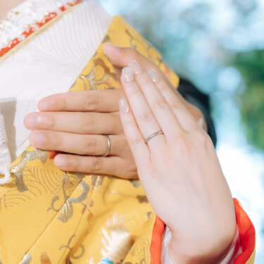 指輪を付けた手を重ねて色打掛の胸元に添えた、新郎新婦の手元アップ。結婚指輪が主役の和装前撮りのカット