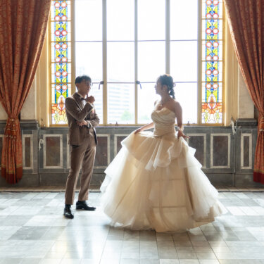光あふれる窓辺で、ドレスを揺らして踊るように回る花嫁を見つめるタキシードの新郎の結婚写真