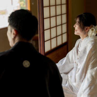 障子の前で新郎の方を向いて微笑む、胡蝶蘭の髪飾りを付けた白無垢の花嫁。和室で撮る和装前撮りの一枚