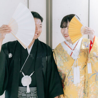 白と金の扇子で顔を半分隠し、手をつないで笑う新郎新婦。心斎橋店のスタジオで撮る和装前撮りの顔隠しポーズ