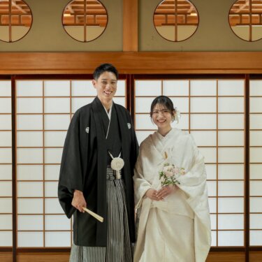 丸い欄間と障子の前に並んで立つ、扇子を持つ紋付袴の新郎とブーケを持つ白無垢の花嫁の和装結婚写真