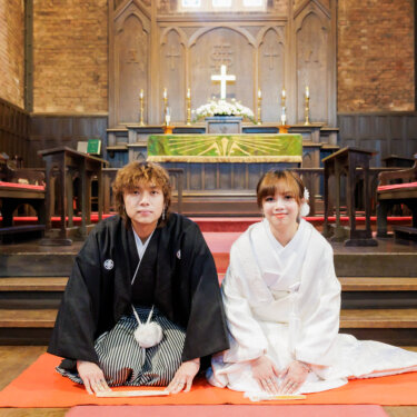 祭壇前の赤い毛氈に並んで正座する紋付袴の新郎と白無垢の花嫁。教会で撮る和装前撮りの、かしこまった正面のカット