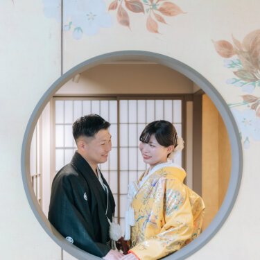 花の描かれた壁の丸窓から、和室で向かい合う紋付の新郎と色打掛の花嫁をのぞく構図。スタジオの和装前撮り