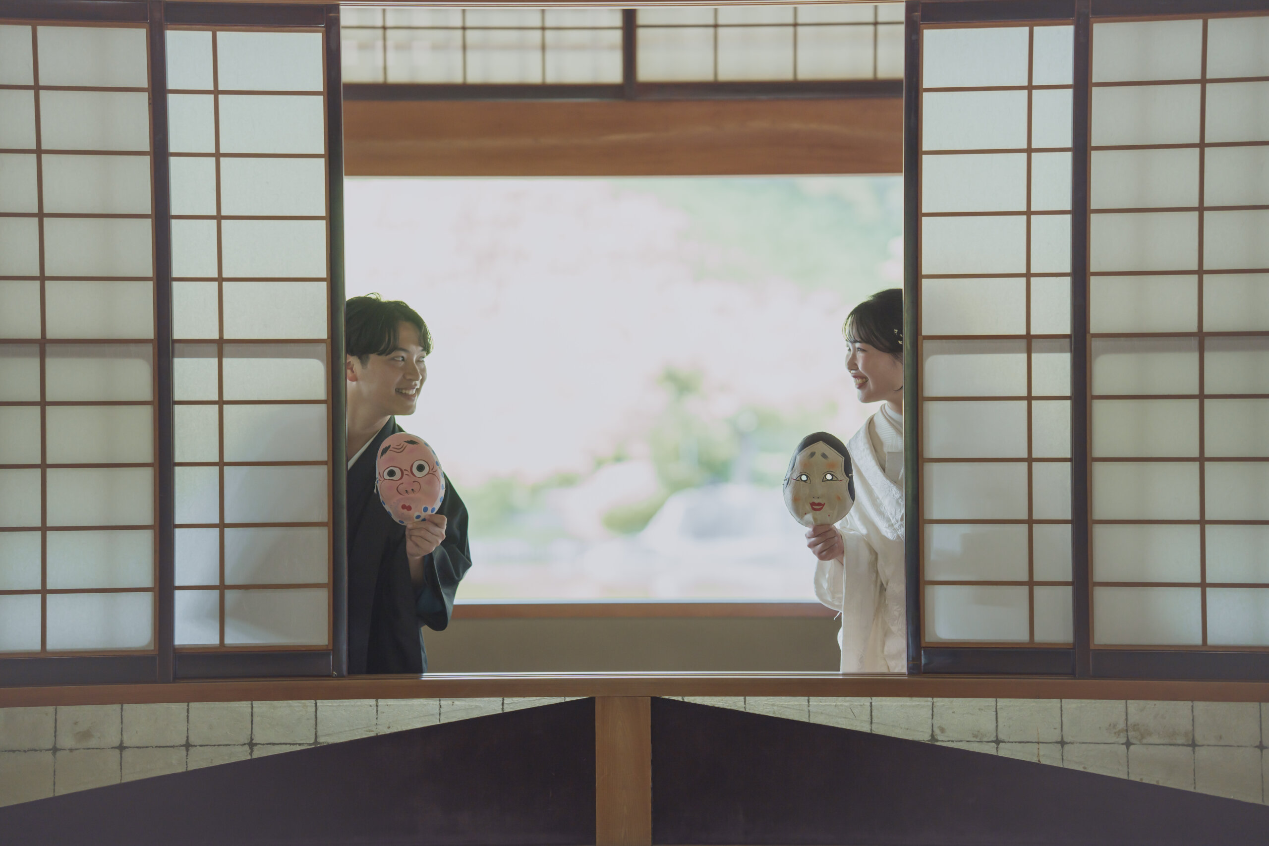 縁側のある和室で、羽織袴の花婿様と白無垢の花嫁様がお面を持ってにこやかに寄り添う写真
