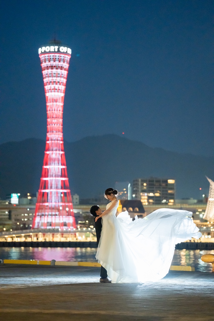 夜空に赤く輝くポートタワーを背に、ドレスの裾を大きく翻す花嫁と寄り添う新郎のナイトフォト前撮り