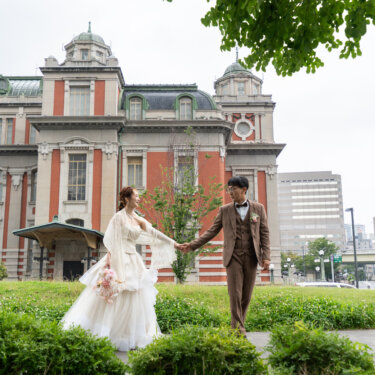 赤レンガの大阪市中央公会堂を背に、芝生の上で手をつないで歩く新郎新婦の中之島ロケーションフォト