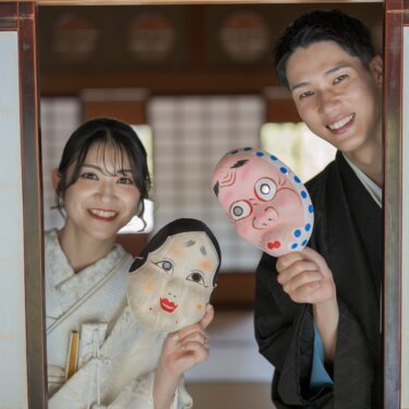 障子の間からおかめとひょっとこのお面を手に顔をのぞかせる新郎新婦。遊び心のある和装前撮りの小物使い