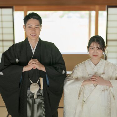 障子の前の和室で手を組んでポーズをとる、紋付袴の新郎と白無垢の花嫁。肩を並べて笑う和装の結婚写真