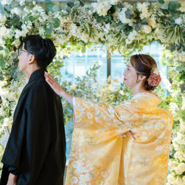 白い花のフラワーアーチの前で、黄色の色打掛の花嫁が黒紋付袴の新郎の肩にそっと手を添える和装前撮り