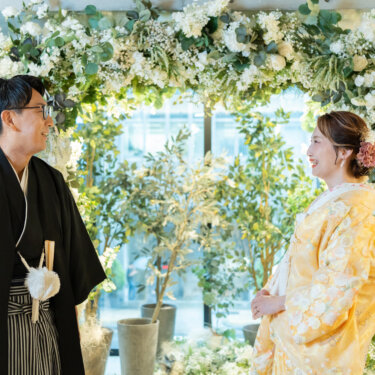 フラワーアーチの前で横を向いて微笑み合う、紋付袴の新郎と黄色い色打掛の花嫁のスタジオフォト