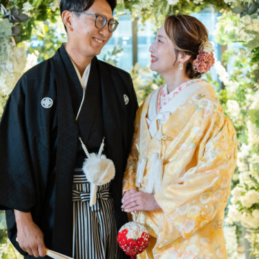 扇子を持つ黒紋付袴の新郎と赤いブーケを手にした黄色の色打掛の花嫁が、笑顔で見つめ合う心斎橋店での和装前撮り