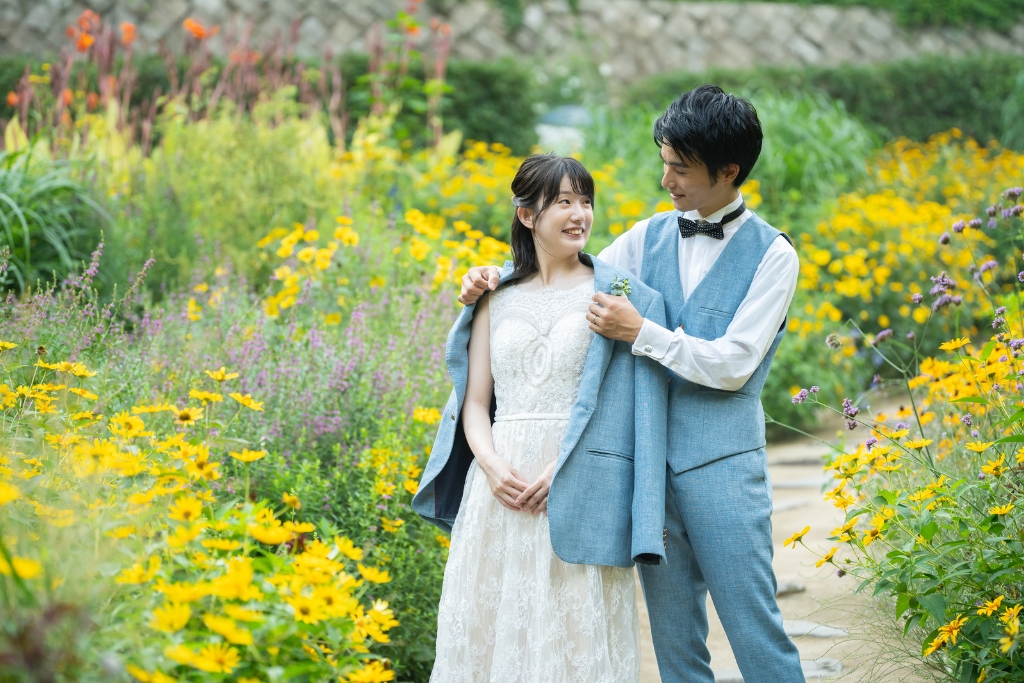 黄色い花が咲くハーブ園の花壇で後ろから肩を抱き寄せる新郎と、振り返って笑う花嫁のナチュラルなフォトウェディング