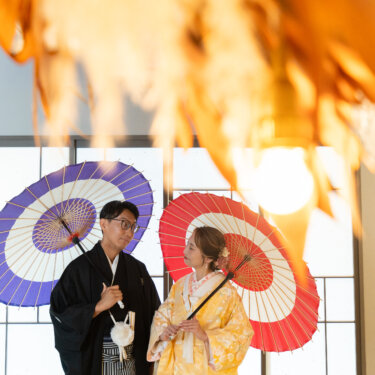 紫と赤の和傘を差して向かい合う紋付袴の新郎と色打掛の花嫁を、手前の草花越しに撮った心斎橋のスタジオでの前撮り