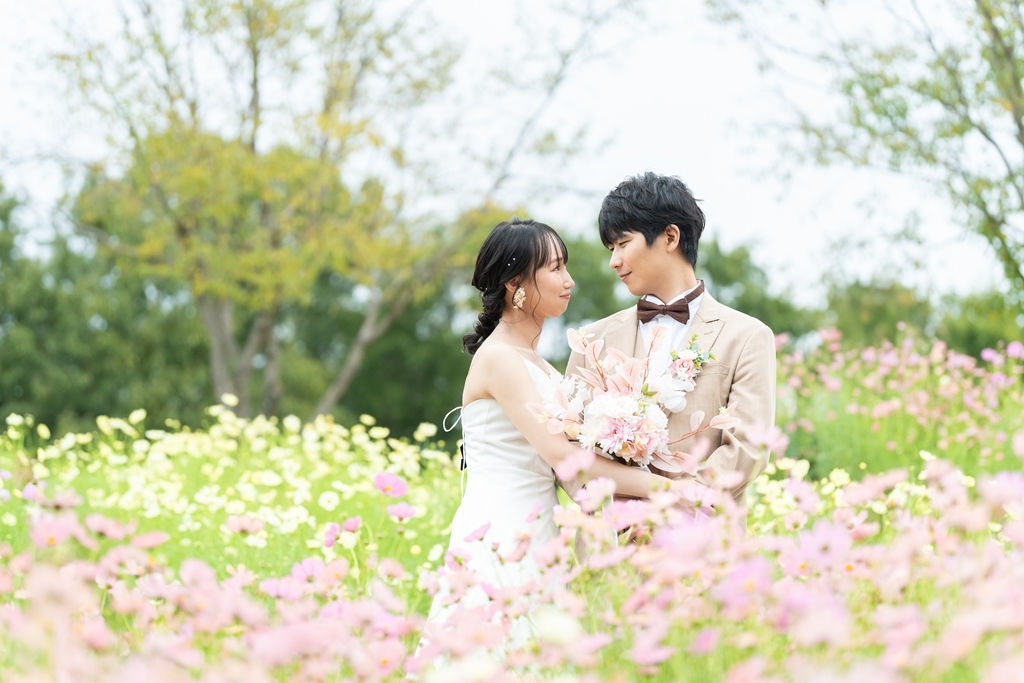 季節の花が一面に咲く丘の中で、花越しに見つめ合うタキシードの新郎とウェディングドレスの花嫁