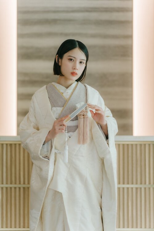 格子と柔らかな照明の和モダンな空間で、扇子とピンクの房飾りを持って正面を向く白無垢の花嫁。白無垢AS105の上半身が分かる和装前撮り