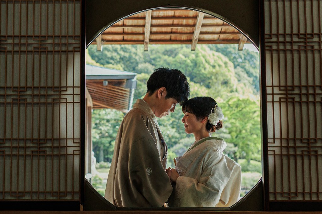 丸い窓と格子の障子に囲まれ、緑豊かな庭を背に額を寄せ合う羽織袴の花婿様と白無垢の花嫁様の写真