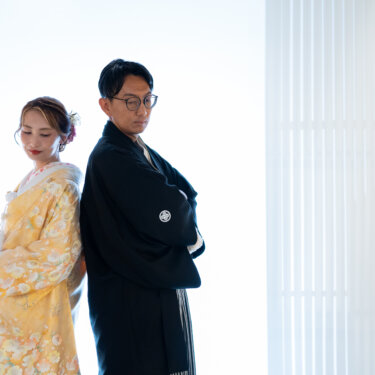 白ホリのスタジオで腕を組み背中合わせに立つ、黒紋付の新郎と色打掛の花嫁のクールな和装前撮りポーズ