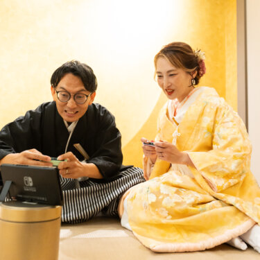 金色の壁の和室セットに並んで座り、ゲームを楽しむ紋付袴の新郎と色打掛の花嫁。ふたりらしさが出る前撮りの一枚