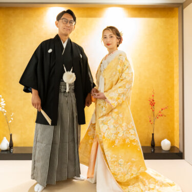 心斎橋店の和室スタジオで、金色の壁の前に並んで立つ紋付袴の新郎と黄色の色打掛の花嫁の和装フォトウェディング