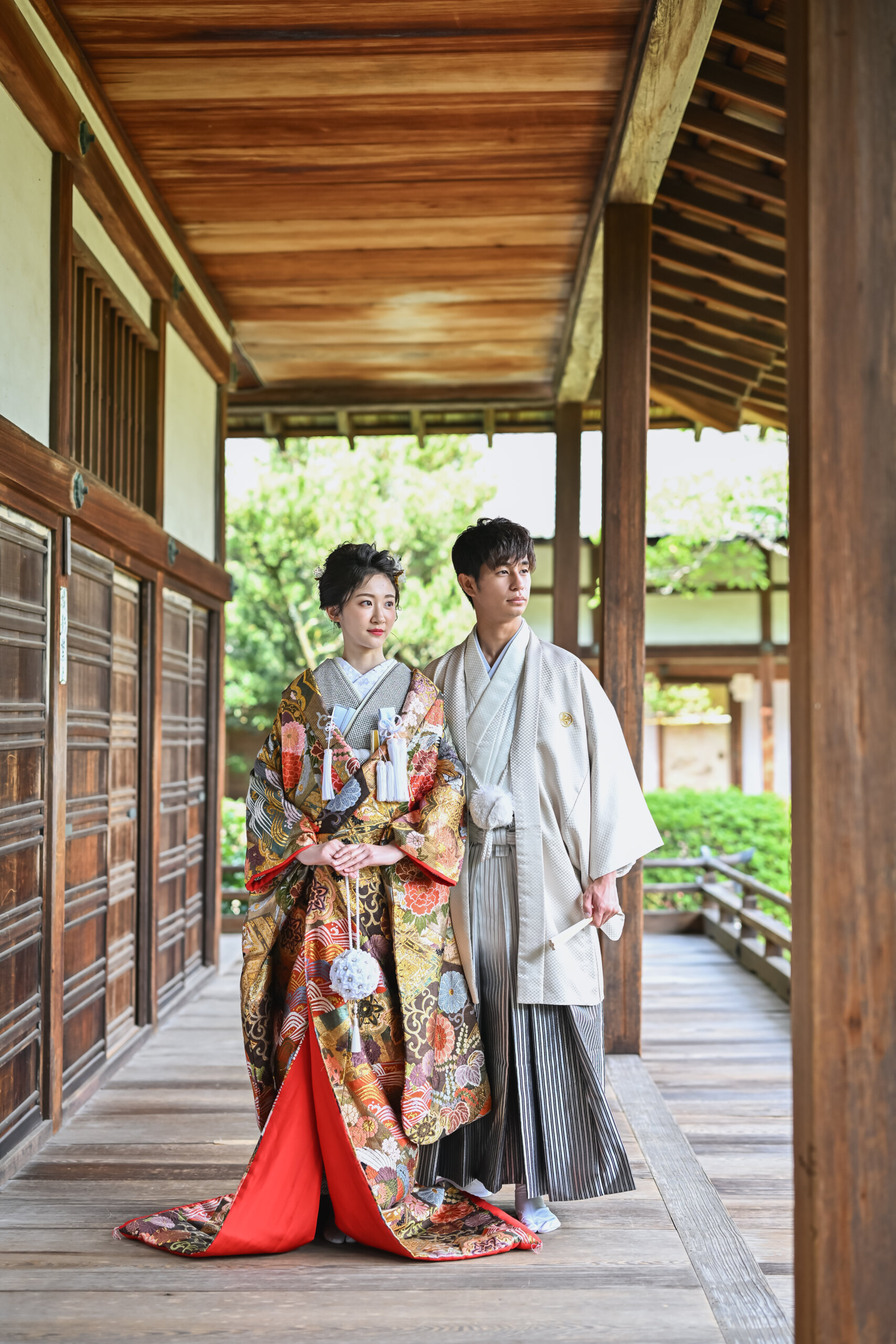 毘沙門堂の木の渡り廊下を並んで歩く、赤い裾の色打掛の花嫁とベージュの羽織袴の新郎の和装前撮り