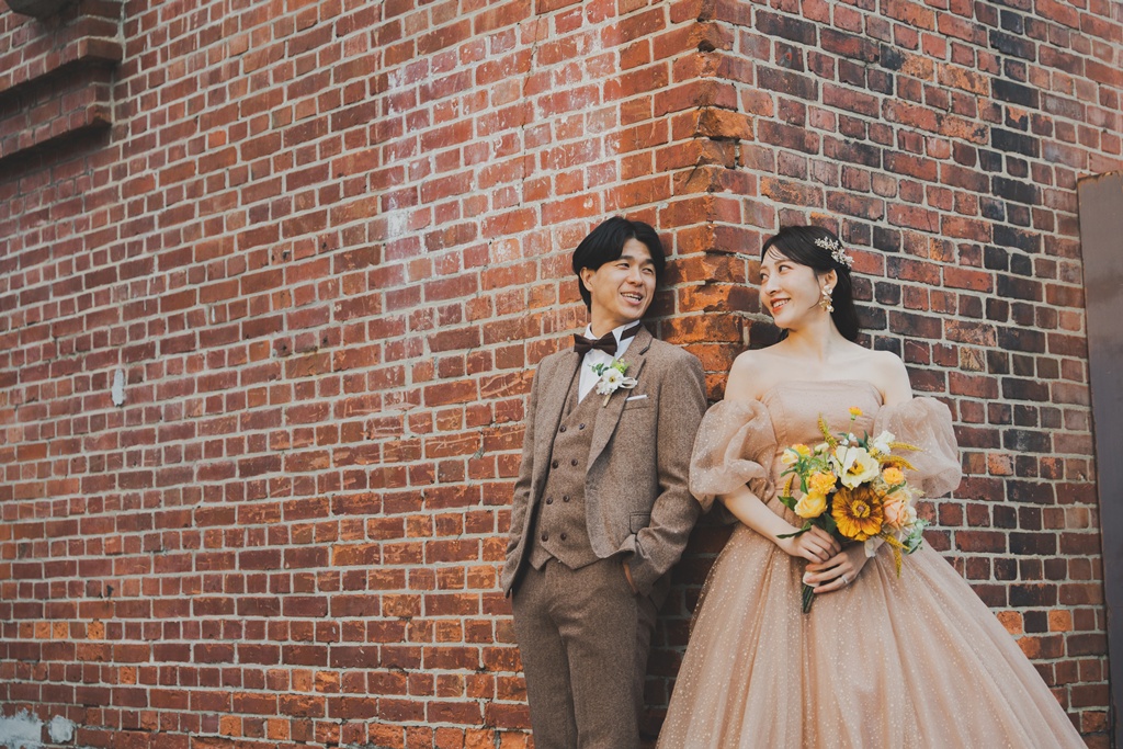 赤レンガの壁にそっともたれ、ブーケを胸元に抱えて微笑むウェディングドレスの花嫁