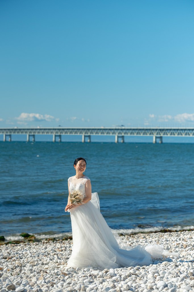 白い小石のビーチで海と大きな橋を背に、ブーケを持って微笑むウェディングドレス姿の花嫁