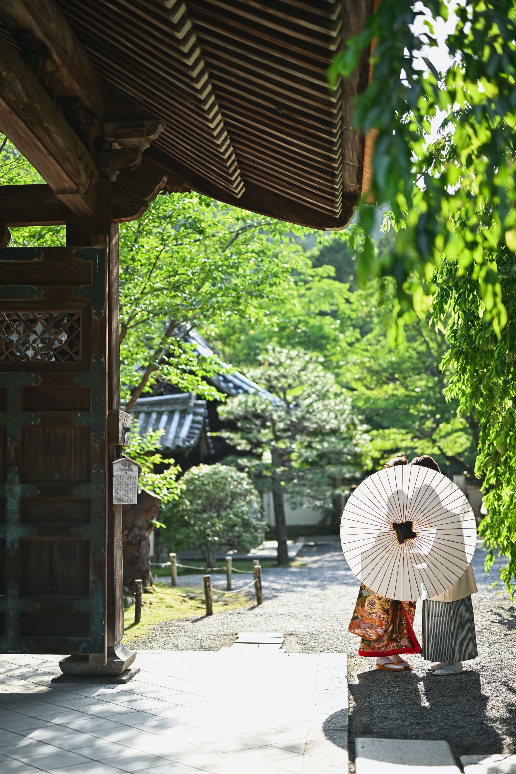 毘沙門堂の門をくぐり、白い和傘を差して新緑の参道を歩く新郎新婦の後ろ姿。京都のお寺で撮る和装前撮り