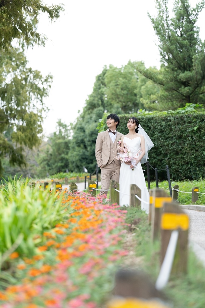 遠くに太陽の塔を望む万博記念公園の緑の中で、寄り添って微笑むタキシードの新郎とウェディングドレスの花嫁