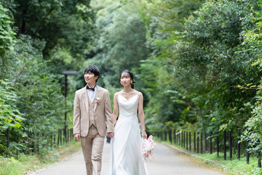 万博記念公園の日本庭園の池のほとりで、緑を背景に向かい合うウェディングドレスの花嫁とタキシードの新郎