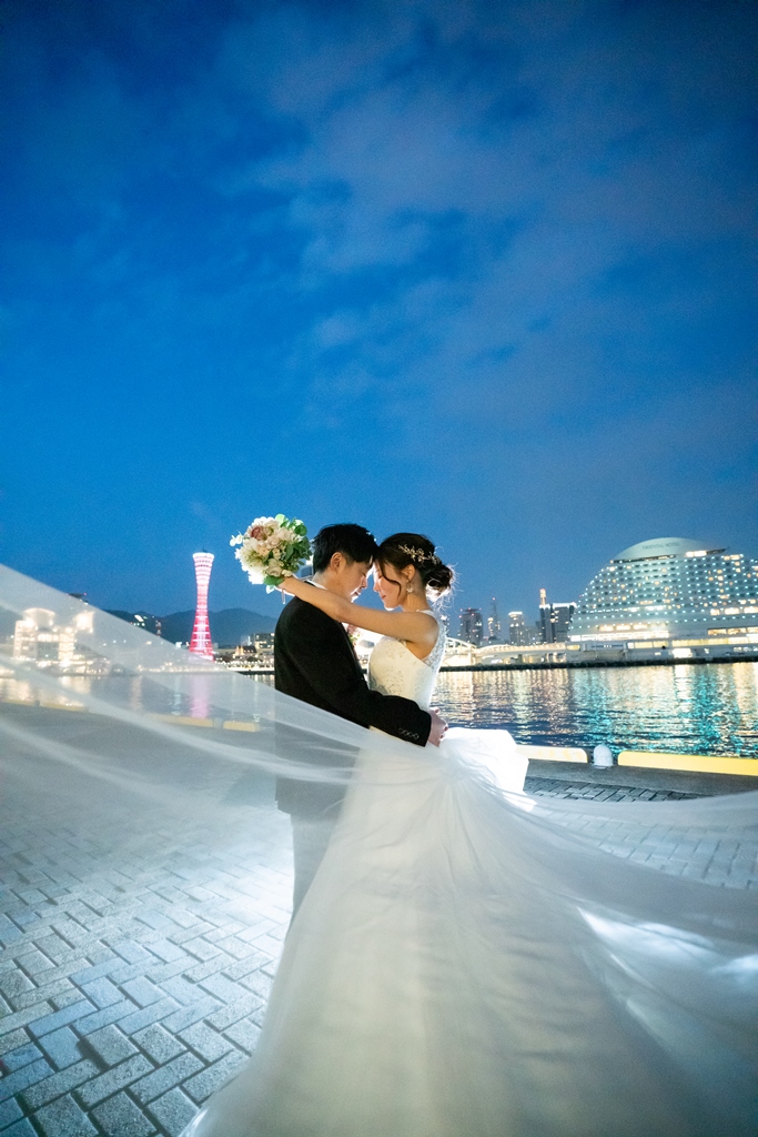 夜の神戸港で神戸ポートタワーを背に、ロングベールをなびかせて抱き合う新郎とウェディングドレスの花嫁。夜景が美しい洋装前撮り