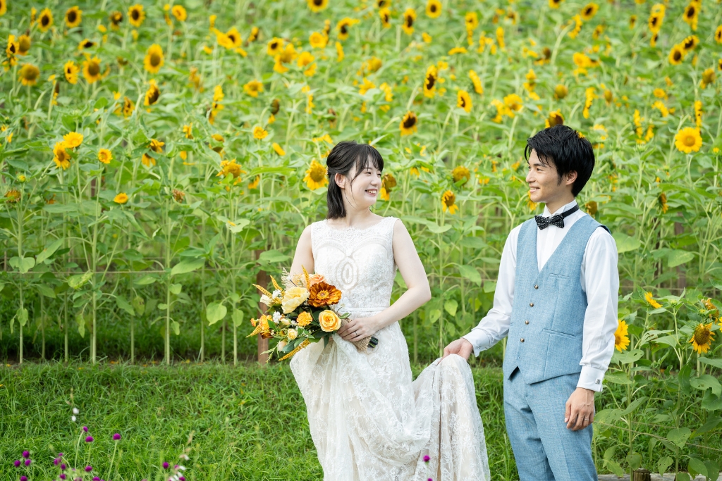 一面のひまわり畑の小道を、ドレスの裾を持ち上げて歩くレースドレスの花嫁とブルーのベストの新郎の夏の前撮り