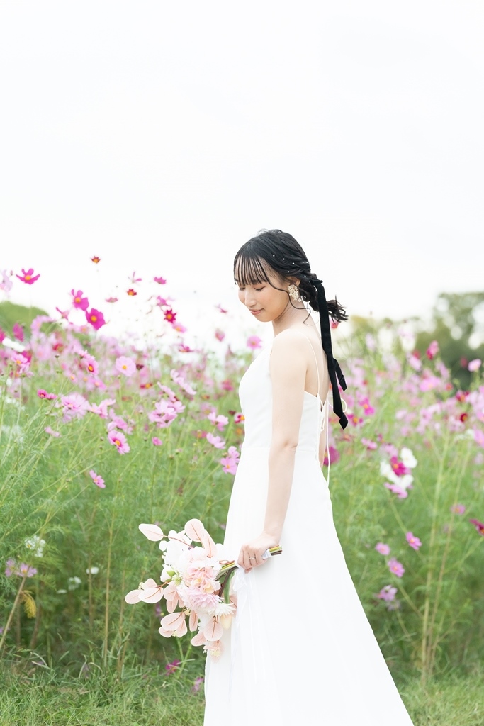 風に揺れる季節の花畑の中で、ブーケを手に振り返って微笑むウェディングドレスの花嫁