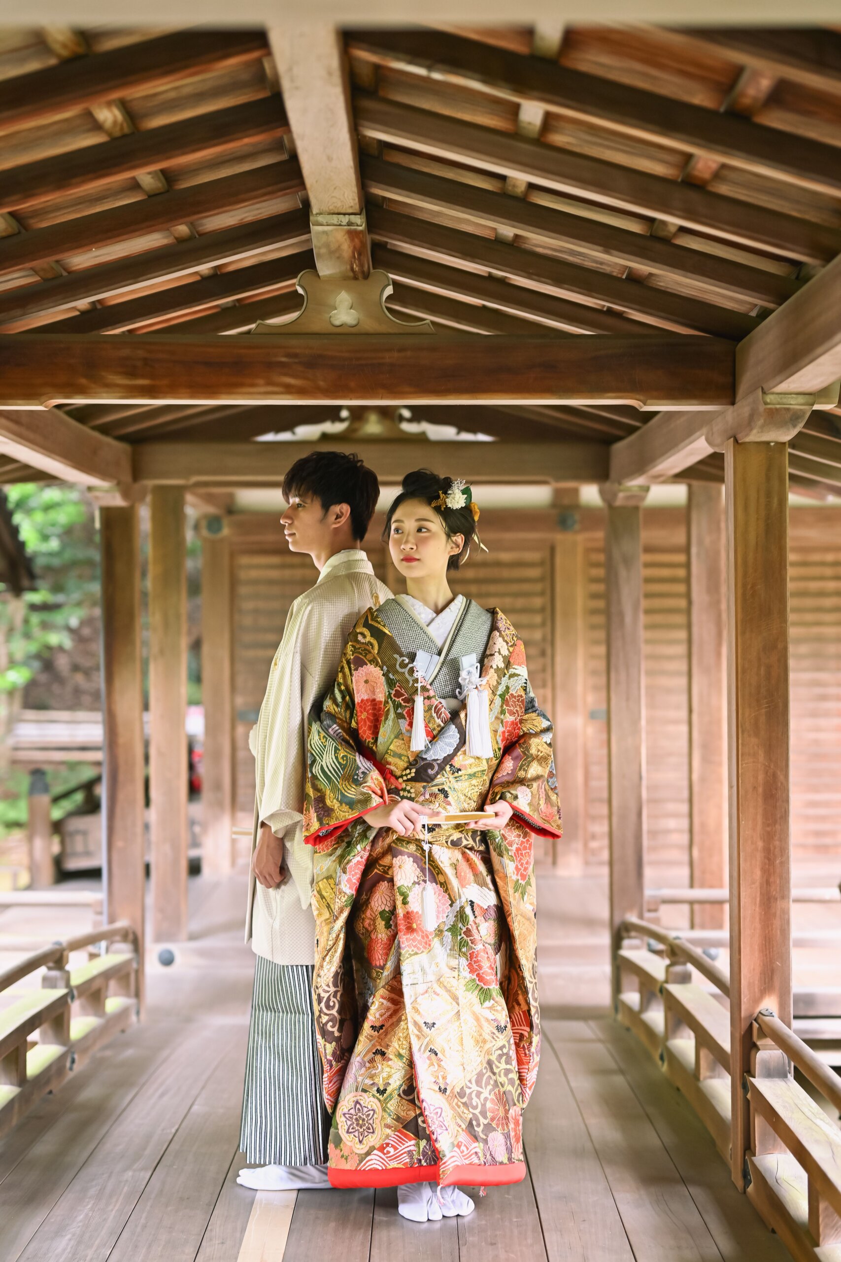 木の天井が美しい渡り廊下で、オレンジ色の色打掛をまとい新郎の前に立つ花嫁のクラシカルな和装前撮り