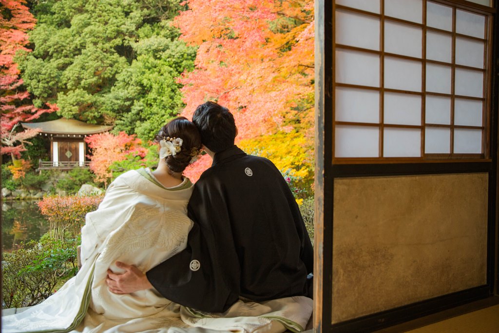 紅葉に染まる毘沙門堂の庭園を望む縁側で、白無垢の花嫁の肩を抱く黒紋付の新郎の後ろ姿。秋の和装前撮り