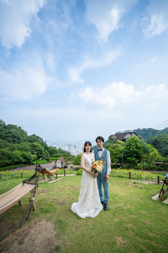 ハンモックのある芝生から神戸の街を見下ろす布引ハーブ園で、青空の下に並ぶ新郎新婦のロケーションフォト