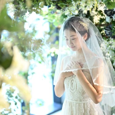 緑の葉越しに、ベールをかぶり胸元で手を重ねてうつむく花嫁。ウェディングドレスの繊細なレースが映える前撮り