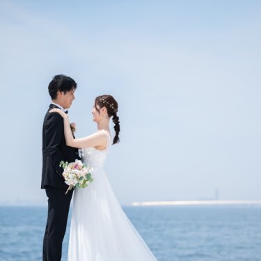 水平線が広がる海を背景に、見つめ合うタキシードの新郎と編み込みヘアの花嫁。爽やかな海辺のウェディングフォト